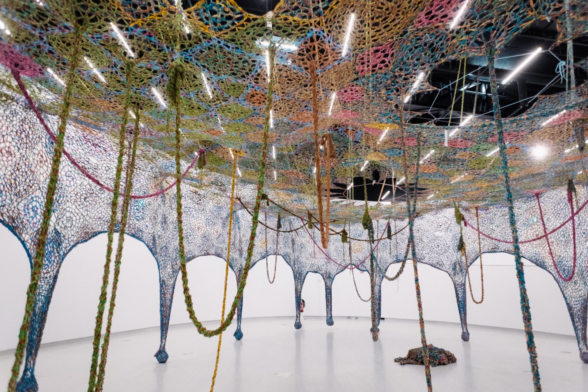 an installation view of an expansive crocheted installation suspended in a large gallery space with rope-like pieces and weights draping it down to the floor