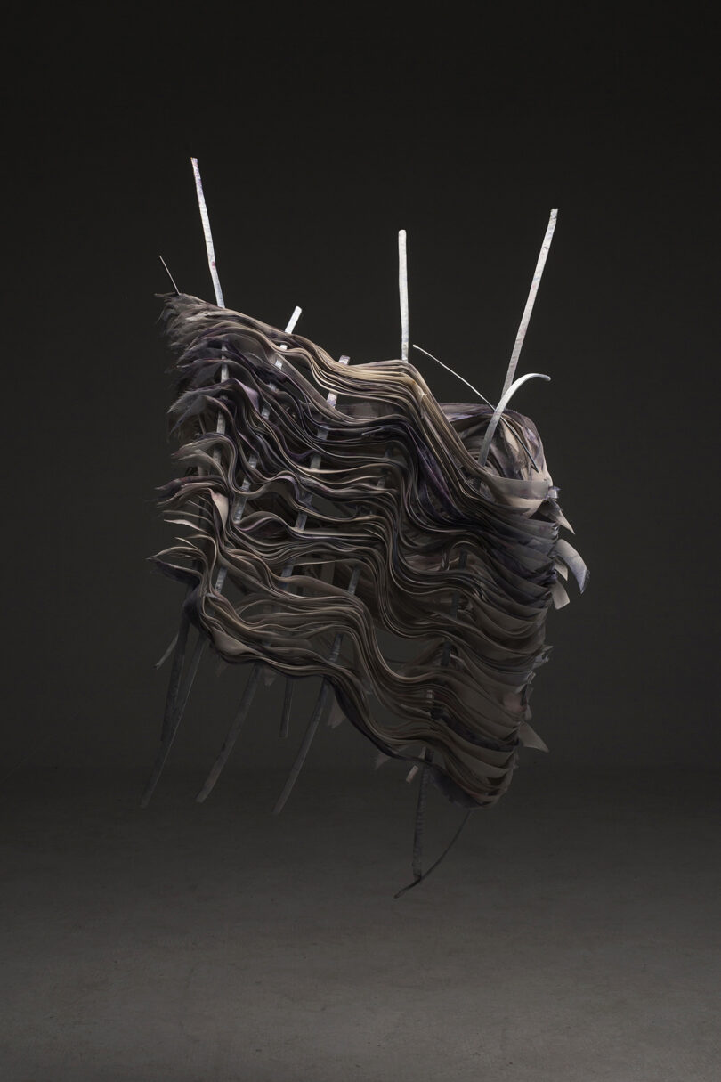 A sculptural artwork featuring layered, undulating dark material with protruding rods, displayed against a gray background.