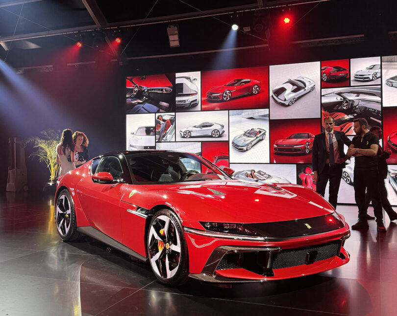 A Ferrari 12Cilindri on display at an automotive event, surrounded by people and large screens showing car designs.