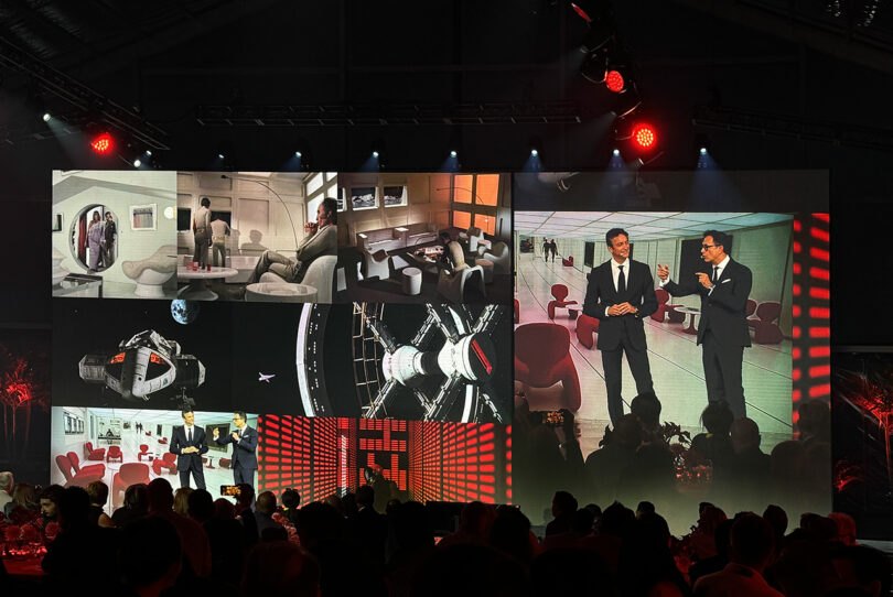 A large event screen displaying a variety of images including astronauts, spacecraft interiors, and two men conversing beside a Ferrari 12Cilindri sports car, viewed by an audience under red lighting.