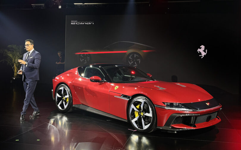 A man in a suit presenting a Ferrari 12Cilindri on stage at a car show, with the Ferrari logo projected in the background.