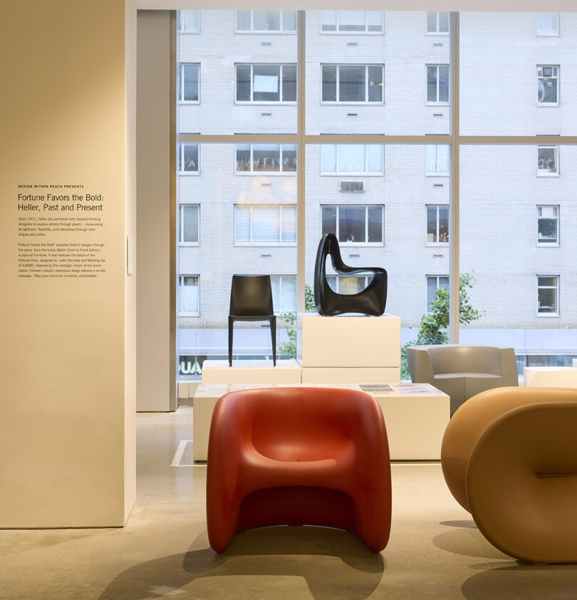 An exhibition room features modern designer chairs, including red, brown, and white pieces, displayed on white podiums in front of large windows with a cityscape view.