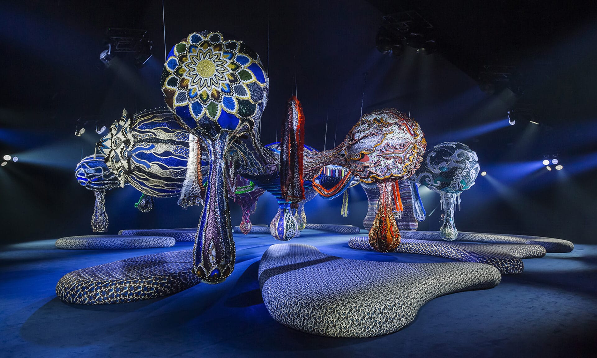 A photograph of a set designed for a fashion show consisting of a raised and rounded runway that slithers through the ground plane of the image while bulbous, multicolor sculptures floating overhead.