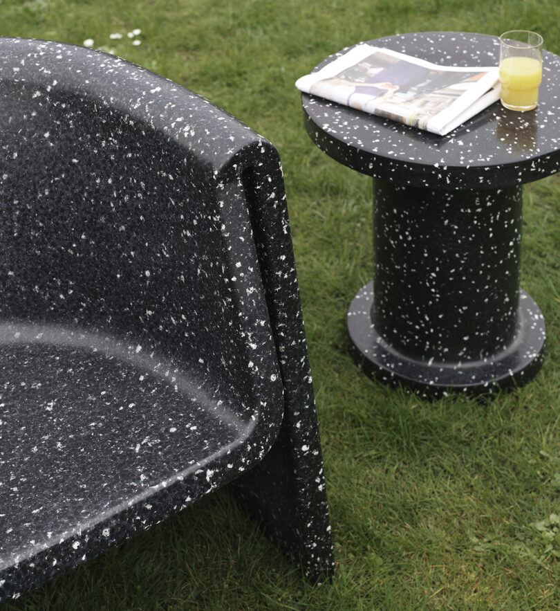Outdoor lounge chair and a side table. The table has a newspaper and a glass of orange juice on it, placed on a grassy lawn.
