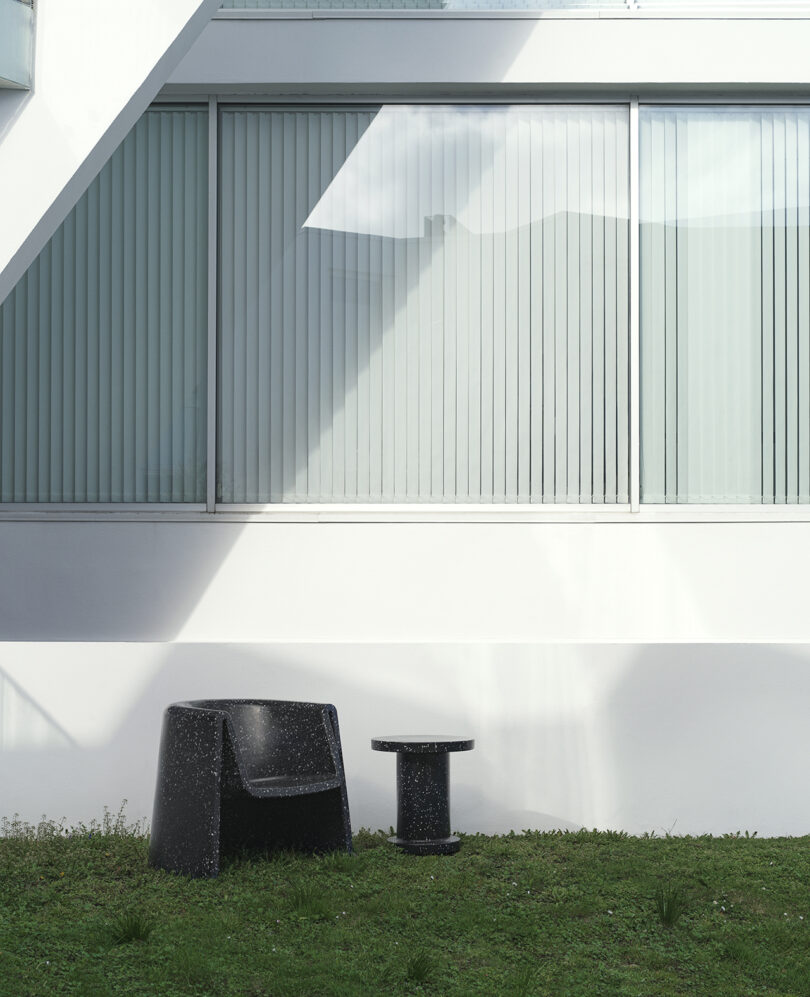 A black lounge chair and matching table sit on grass in front of a building with large windows featuring vertical blinds.