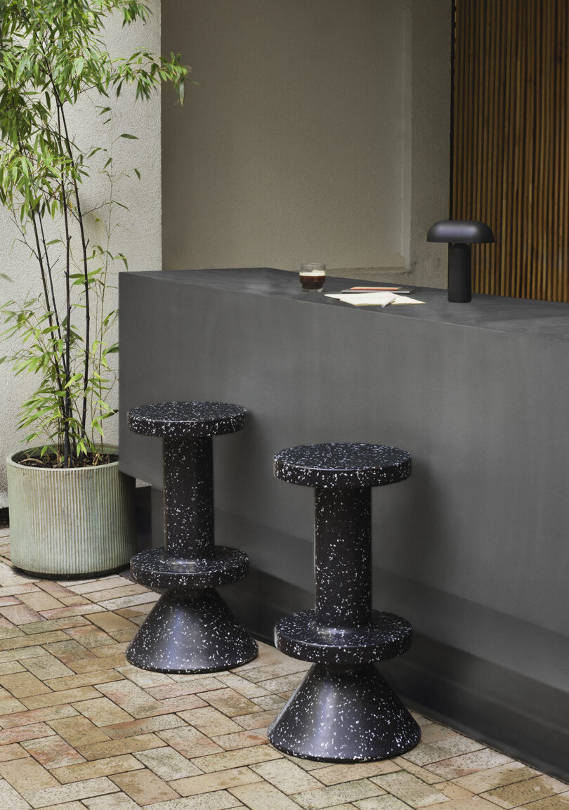 Two round black barstools with white speckles stand by a gray counter. A drink and a book rest on the counter beside a black lamp. A potted plant is on the left of the scene.