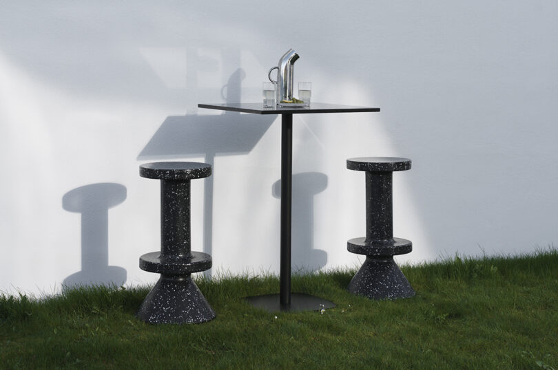 Two black speckled barstools and a square cafe table with two glasses and a pitcher are placed on a green lawn against a white wall.