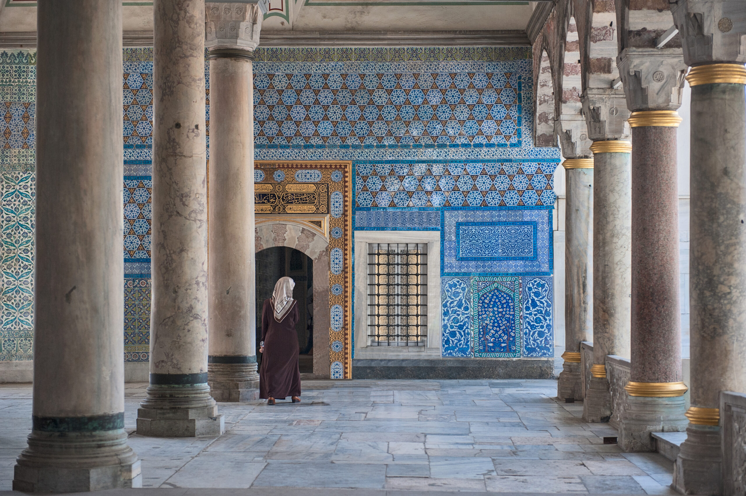A woman walks towards a doorway in a wall covered with intricately patterned blue tiles at Topkapı Palace. The room has several tall, marble columns.