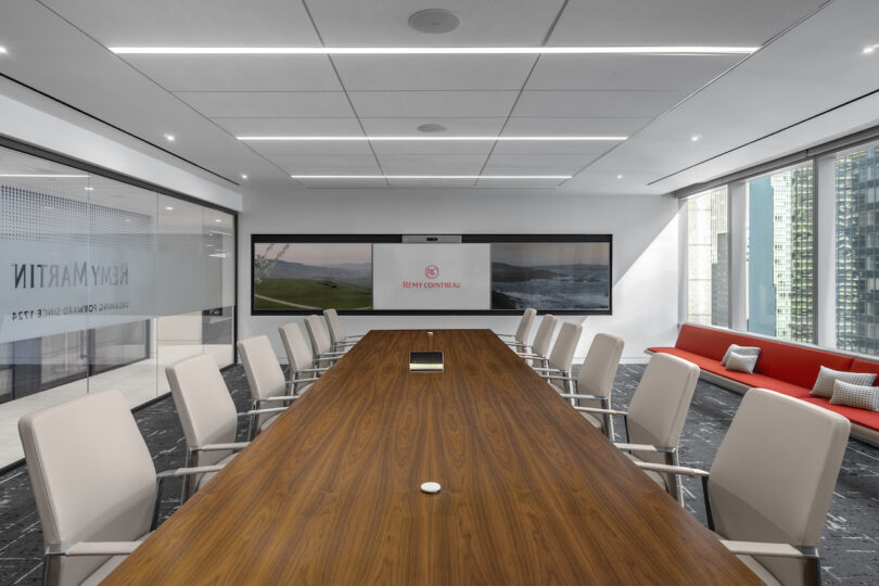 A modern conference room with a long wooden table, white chairs, a large screen displaying a logo, and floor-to-ceiling windows. A red couch with pillows is along one wall