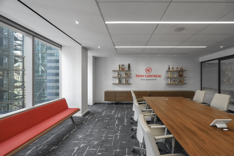 A modern office meeting room with a long wooden table, white chairs, a red bench, and a Remy Cointreau logo on the wall. Large windows provide a view of city buildings