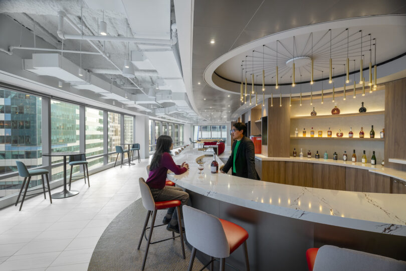 Two individuals are conversing at a modern, curved bar in a spacious, well-lit office lounge with large windows and an array of liquor bottles displayed on illuminated shelves