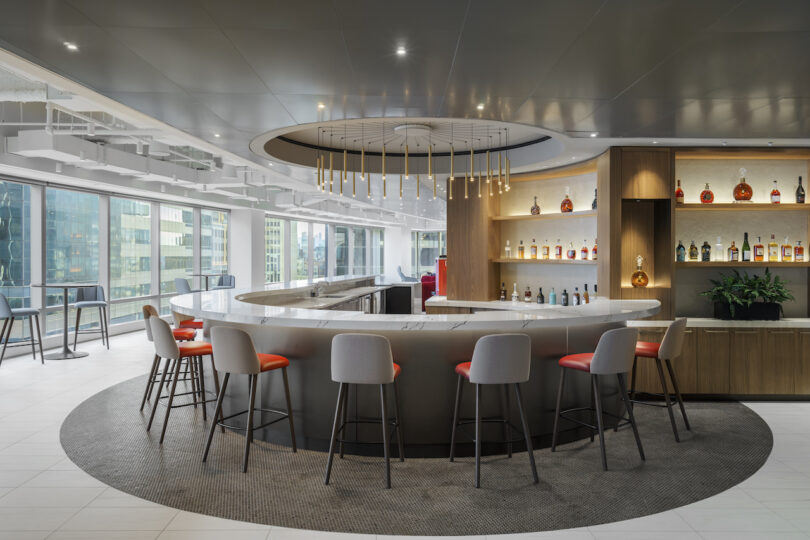 Modern bar area with a circular counter, red cushioned stools, and shelves of bottles against a backdrop of large windows overlooking city buildings. Ceiling features a contemporary light fixture