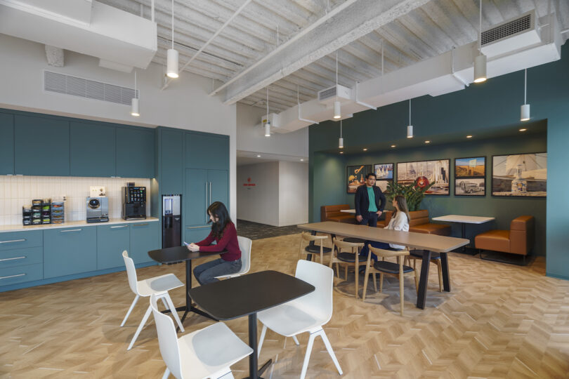 A modern break room with a kitchenette on the left, tables and chairs in the center, and a seating area with artwork on the wall in the background. Three people are present, one seated and two standing