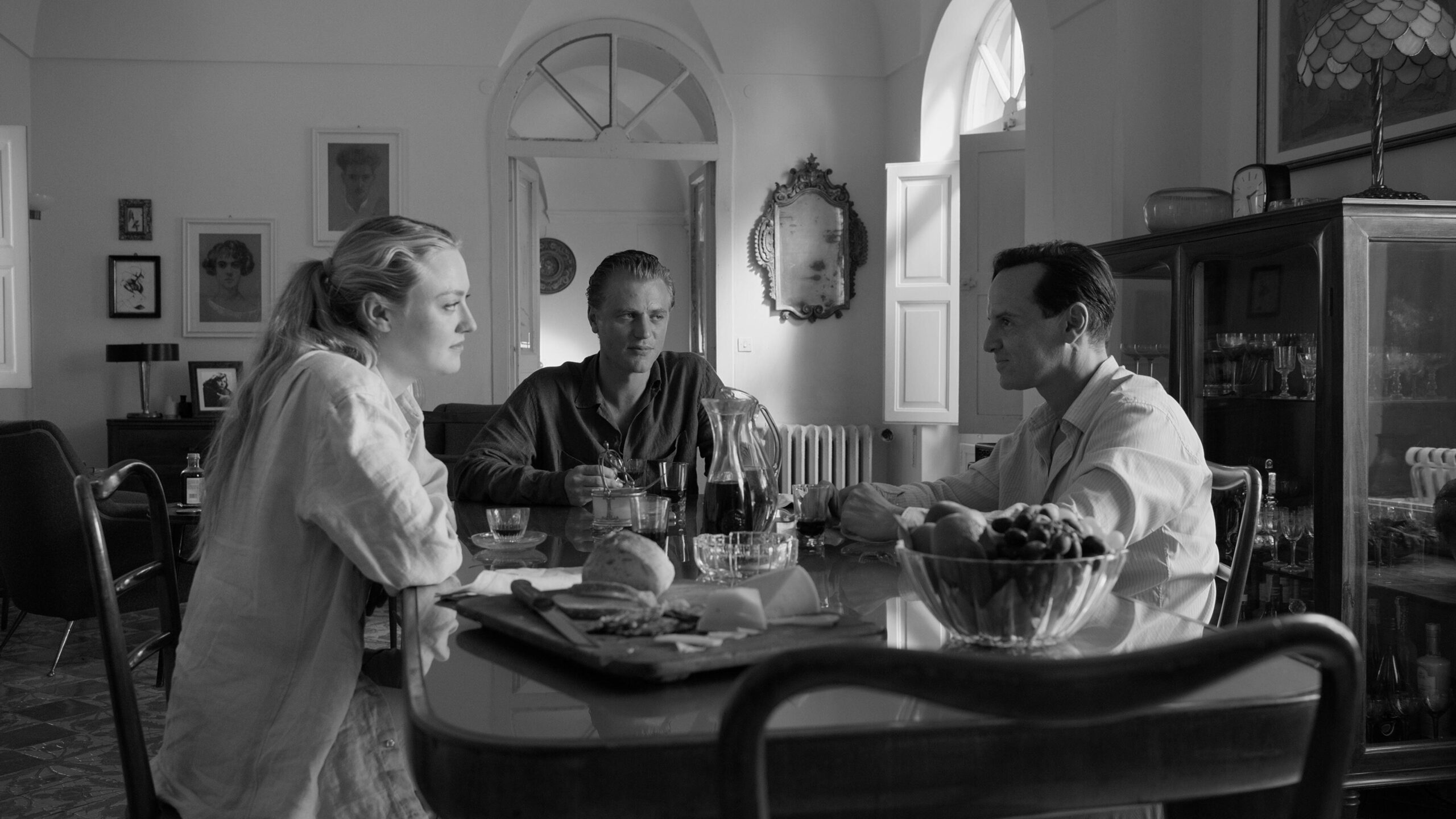 Black-and-white still from a movie scene where two men and a woman are sat a table together. The man and woman look suspiciously at the third man
