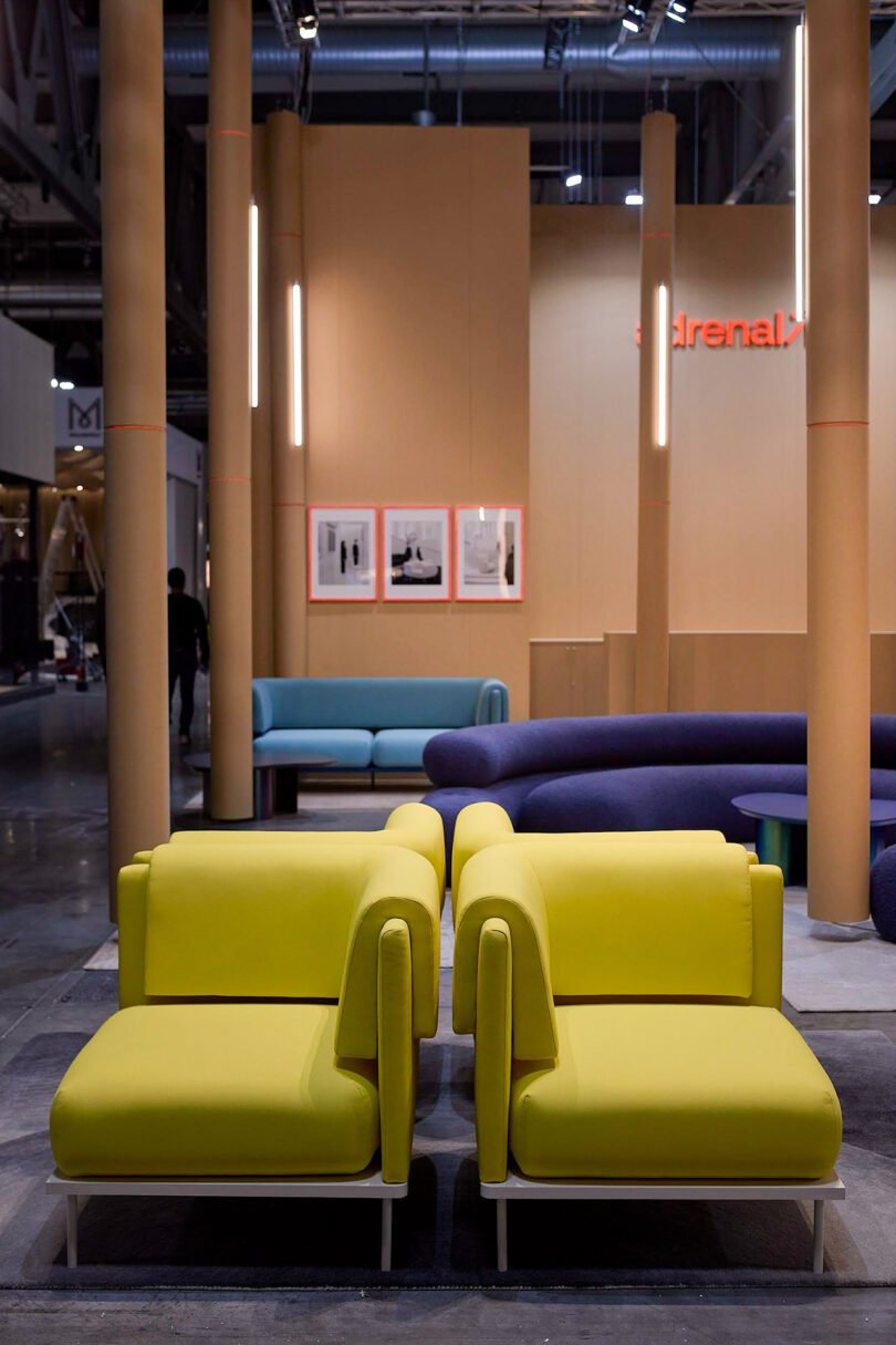 A modern showroom features yellow modular seating in the foreground, blue seating in the background, and framed artworks on a beige wall under industrial lighting.