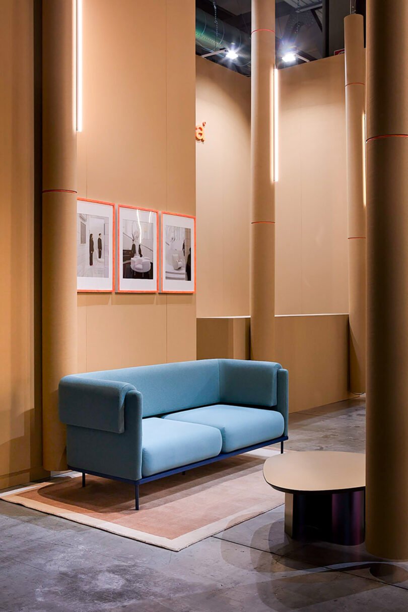 A modern showroom with a light blue sofa, round coffee table, beige walls, framed pictures, and vertical light fixtures.