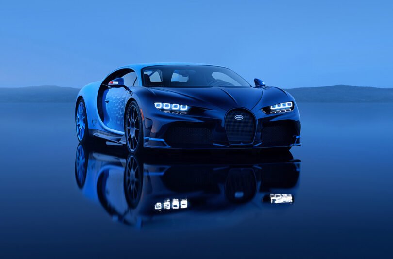 A sleek, black Bugatti Chiron with illuminated headlights is parked on a reflective surface against a blue-tinted background.