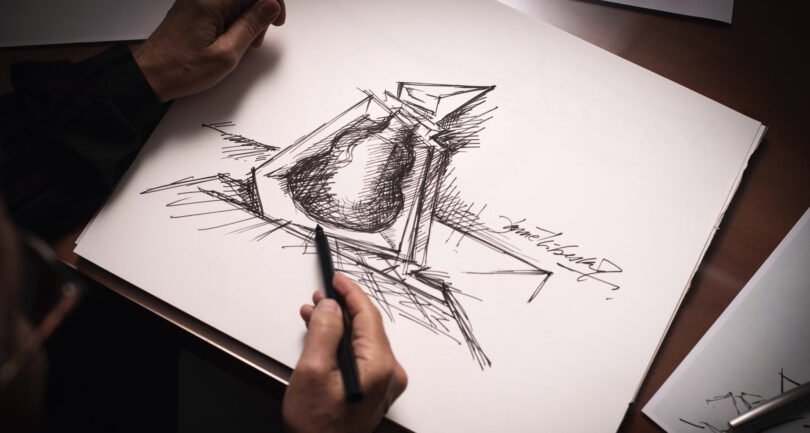 A person sketches an abstract design of a cognac bottle on white paper using a pen, their hand, holding the pen, in the foreground.