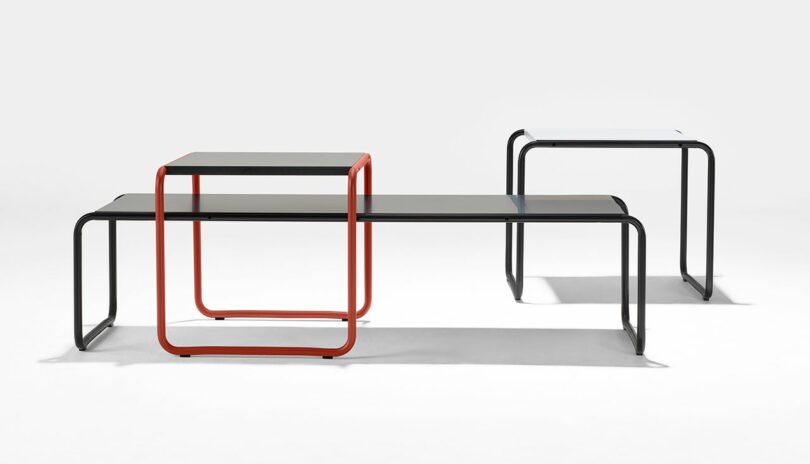 Three modern nesting tables: a red-bordered table on top of a larger black table, next to a separate smaller black table, all with minimalist metal frames and flat surfaces.