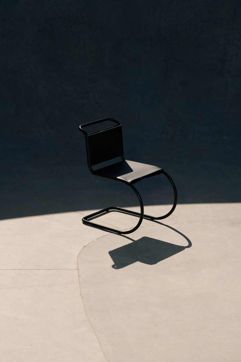 A modern black chair with a curved metal frame casting a shadow on a smooth concrete surface, partially illuminated by sunlight.
