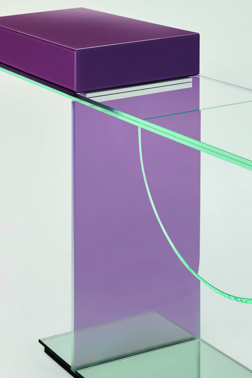Detail of a modern glass console featuring a purple rectangular top, a vertical support, and a curved glass shelf.