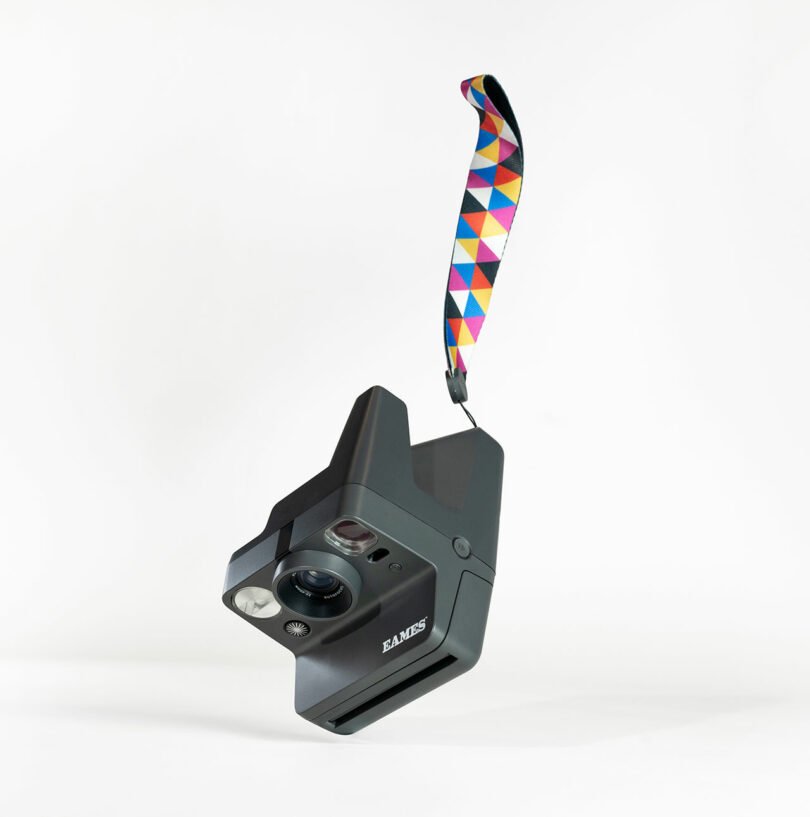 A black instant camera with a geometric-patterned strap is suspended in mid-air against a plain white background.