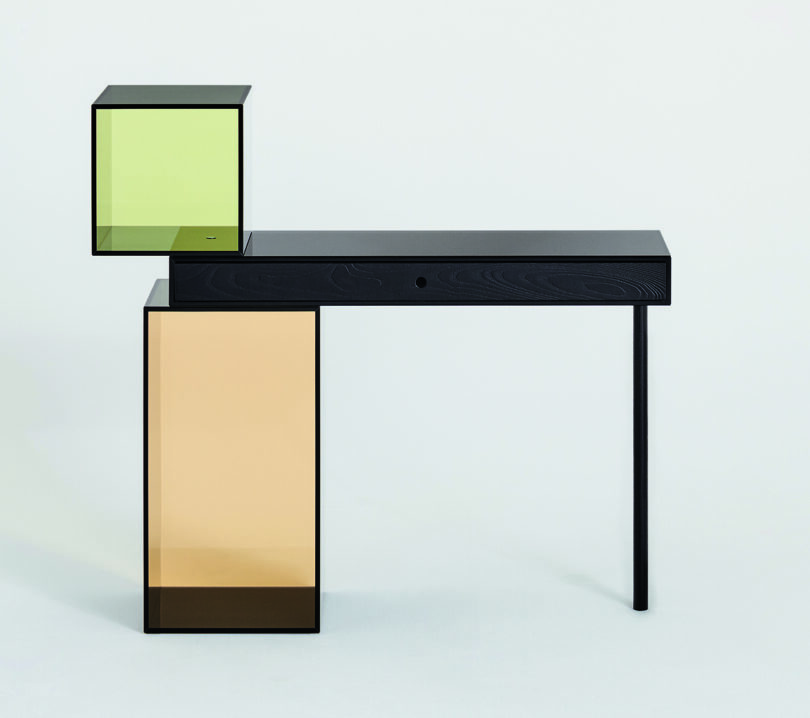 A modern, minimalist desk with an asymmetrical design featuring black surfaces and transparent colored panels in green and orange. One leg is black while the other supports a green cube.