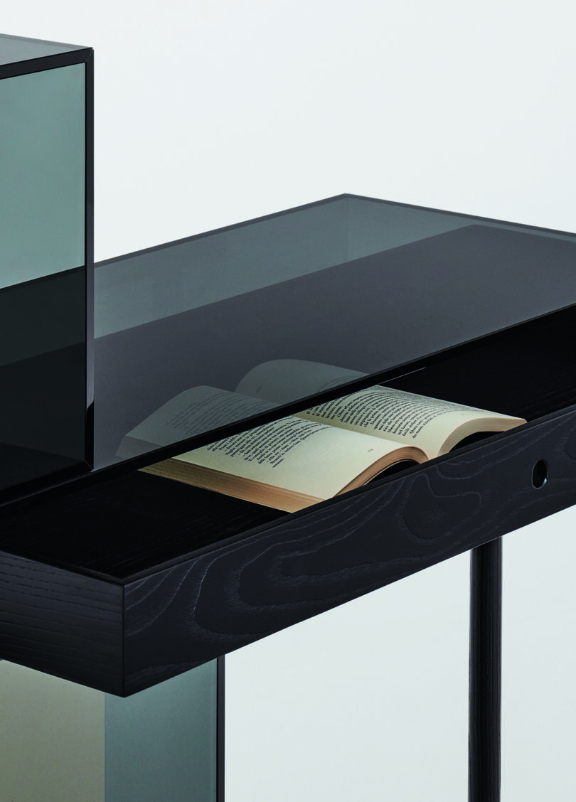 Detail of a modern, minimalist desk with an asymmetrical design featuring black surfaces and transparent colored panels in green and orange. One leg is black while the other supports a green cube.