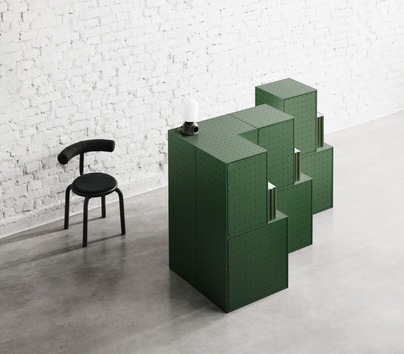 A modular, green storage unit from the String Furniture X FUWL Center collection is composed of blocks and is placed near a minimalist black chair in front of a white brick wall in a modern, sparsely furnished workspace.