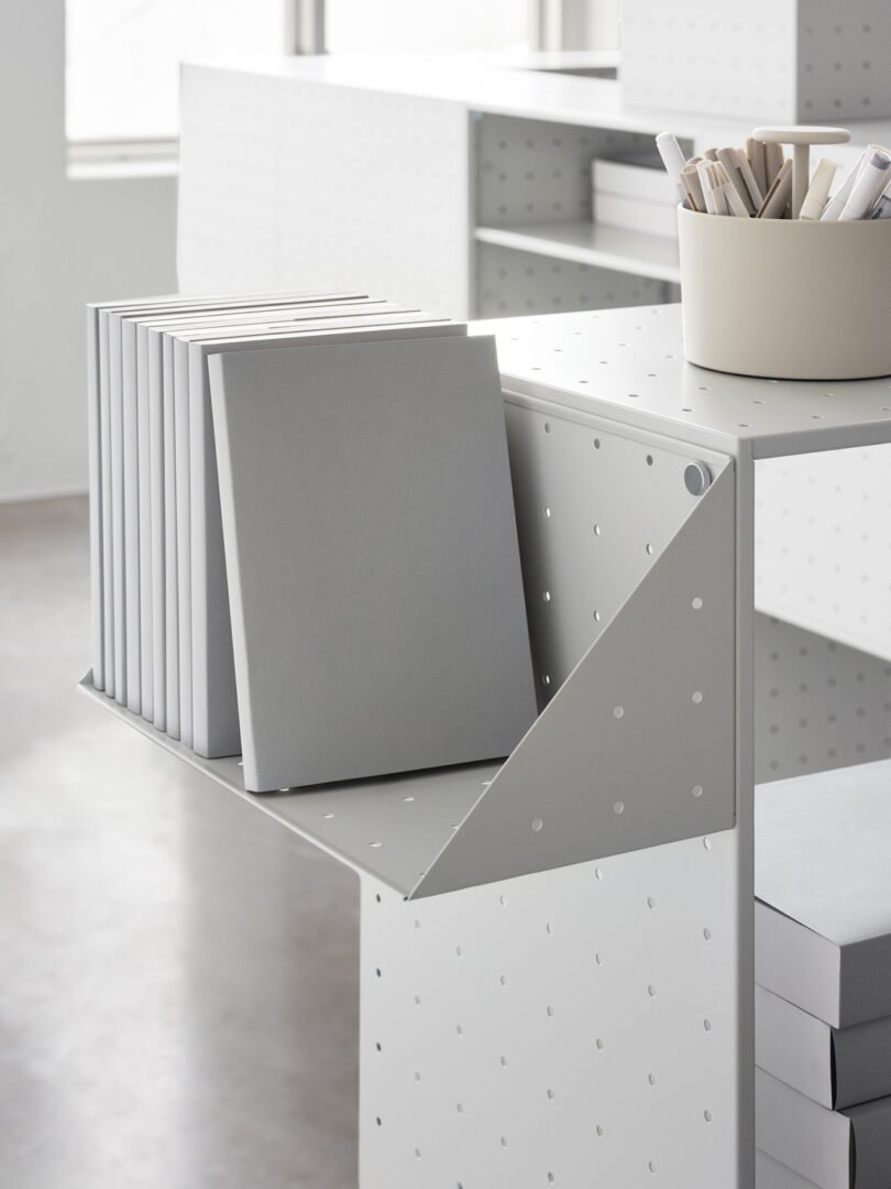 A modern, minimalist bookshelf from String Furniture X FUWL Center with several gray books neatly arranged on it. A round container holding rolled-up paper is placed on the top shelf, making it an ideal piece for workspace furniture.