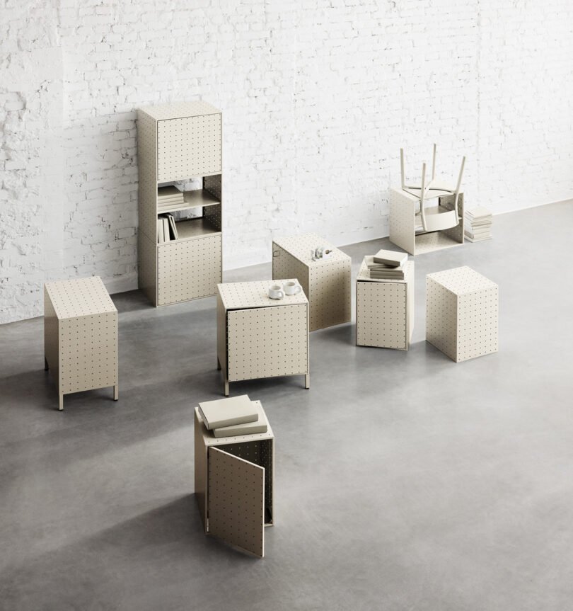A minimalist room with white brick walls features various beige modular String Furniture pieces, including tables, chairs, and storage units, all arranged on a gray concrete floor.