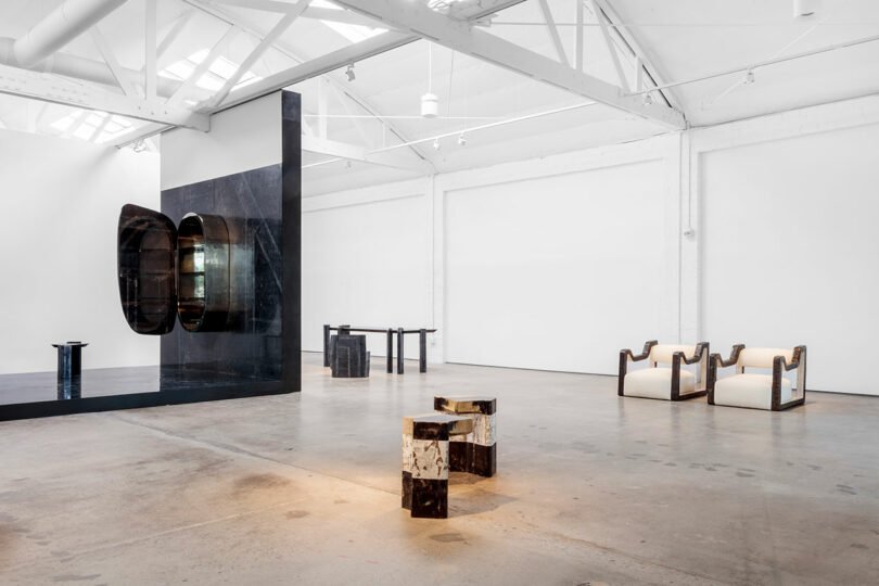 Spacious art gallery with modern, minimalist black and white furniture designed by Vincenzo De Cotiis, featuring abstract sculptures and large white walls under a high, white ceiling with exposed beams.