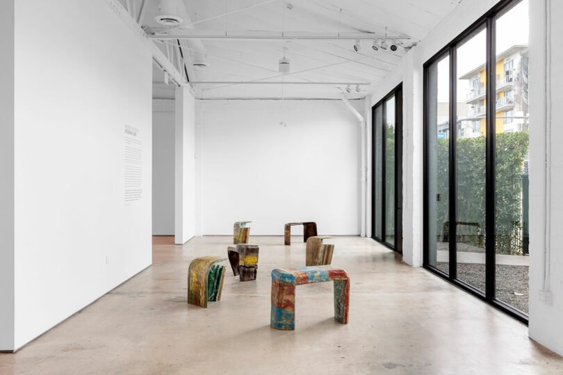 Modern art gallery featuring colorful, abstract sculptures made from various materials, displayed on a polished concrete floor. Large windows line the right wall, letting in natural light. The gallery offers an elegant yet raw aesthetic that captivates visitors.