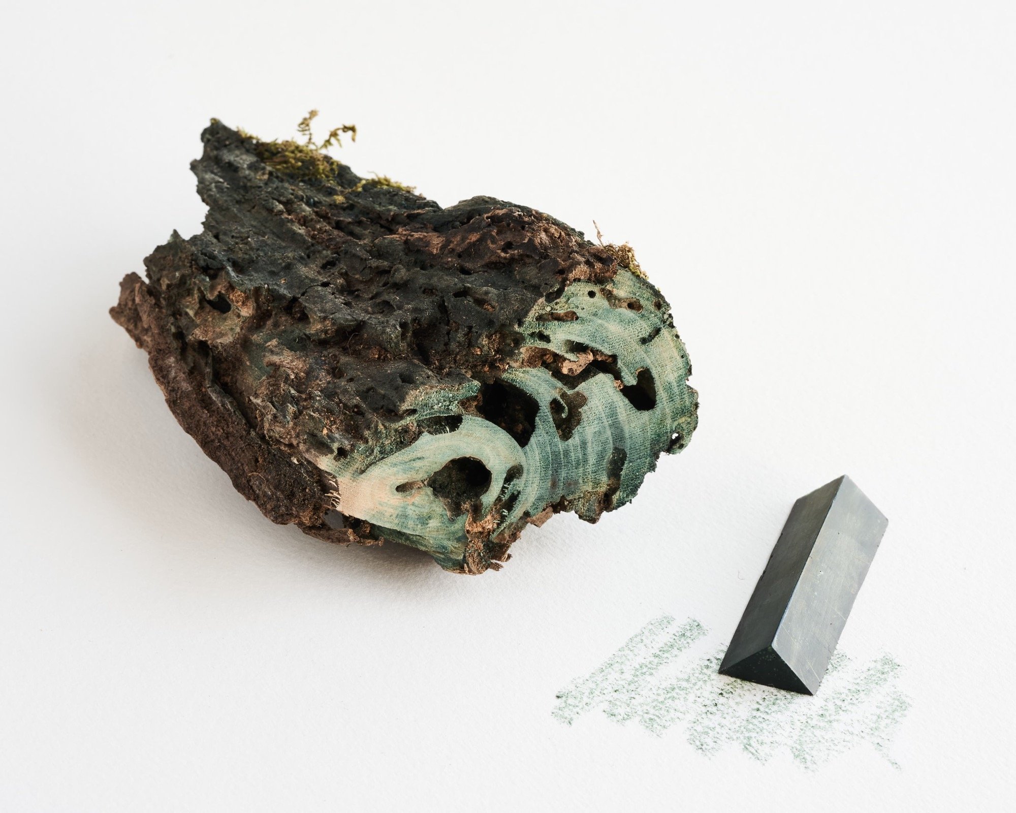 a triangular crayon with a blueish hue, shown next to a piece of timber from which its pigment was ground