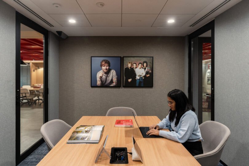 A woman works on a laptop at a conference table in a modern office meeting room with framed photos on the wall and various items on the table