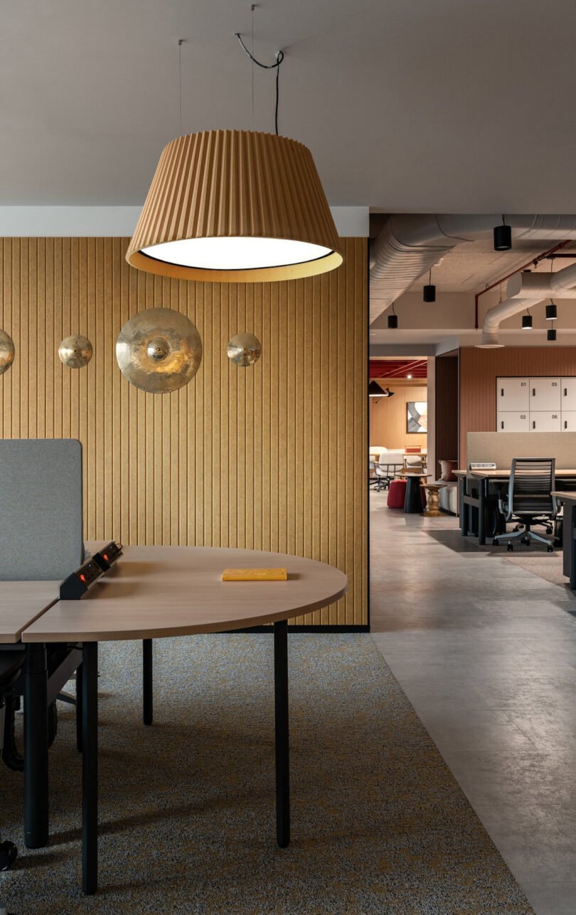 Modern office space with wooden paneling, round tables, and pendant lights. Several workstations and chairs are visible, with a mix of open and secluded seating areas