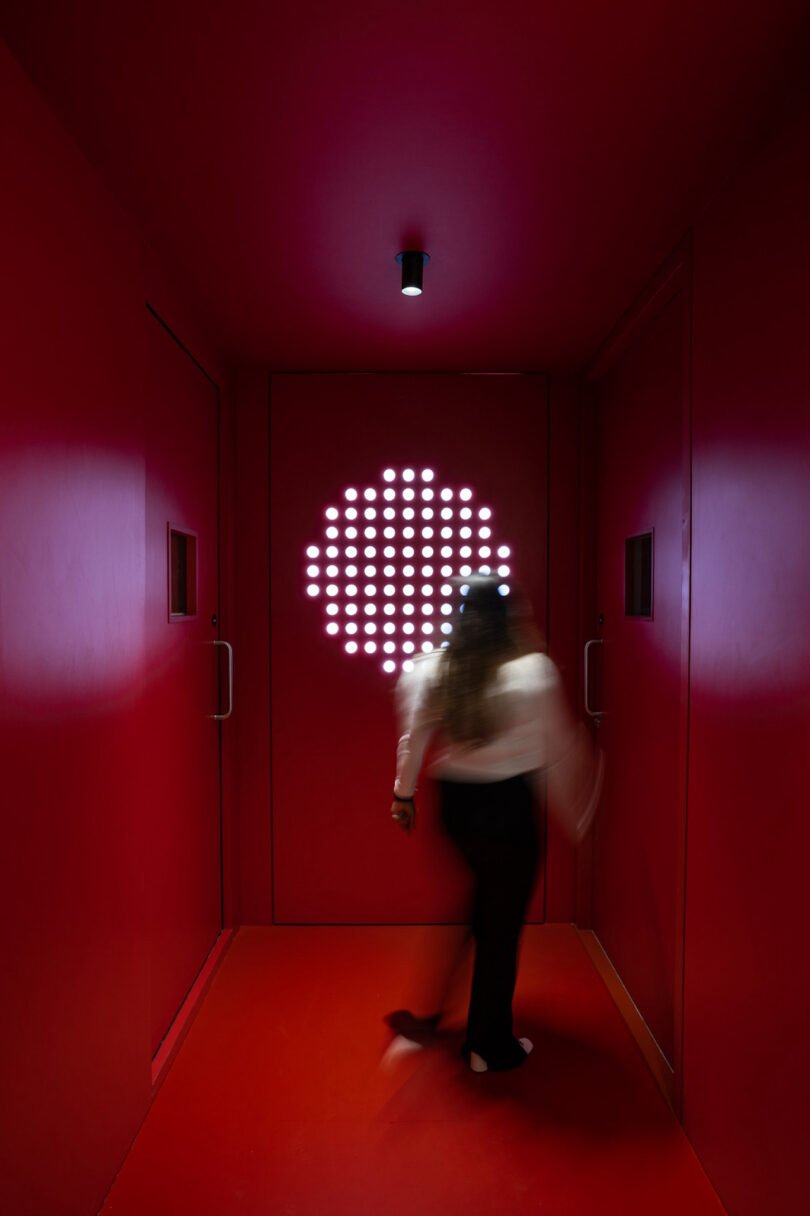 A person stands in a red-lit hallway with white round lights arranged in a circular pattern on the far wall. The person is blurred, indicating motion
