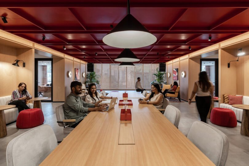 People are working in a modern office space with wooden furniture, red accents, and overhead lighting. Several individuals are seated at a long table with laptops, while others move about or sit in the background