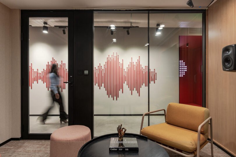 A modern office interior with a glass partition featuring a red soundwave design. A person is walking through a door on the left, and a seating area with a chair and ottoman is in the foreground