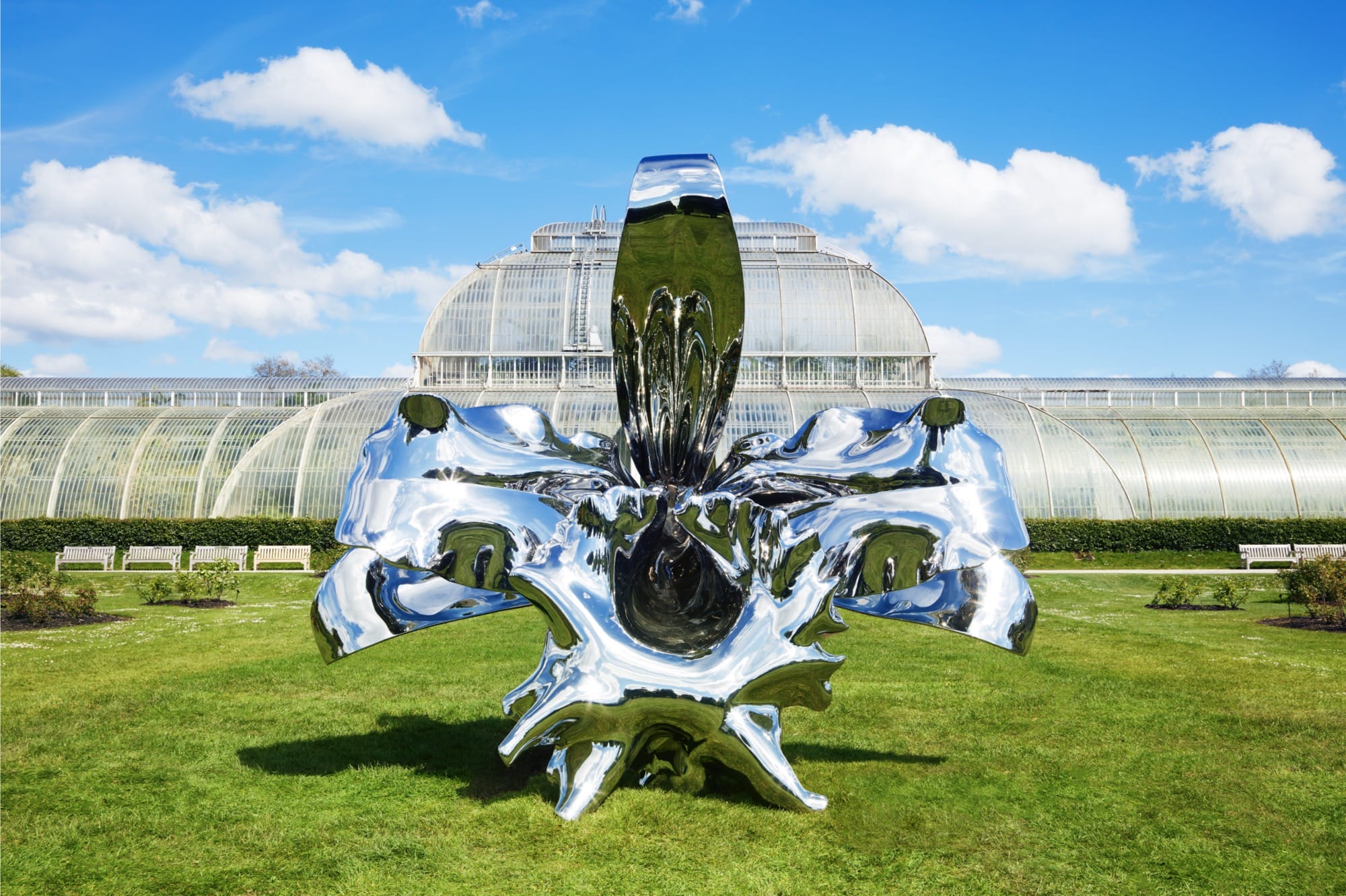 a larger-than-life orchid sculpture coated with chrome, reflecting the sun and sky on the green lawn in front of one of Kew Gardens' greenhouses