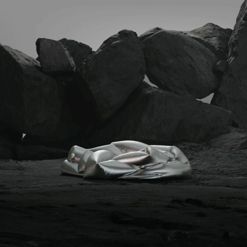 A metallic, inflated sofa lies on a rocky terrain with large boulders in the background under a gray sky