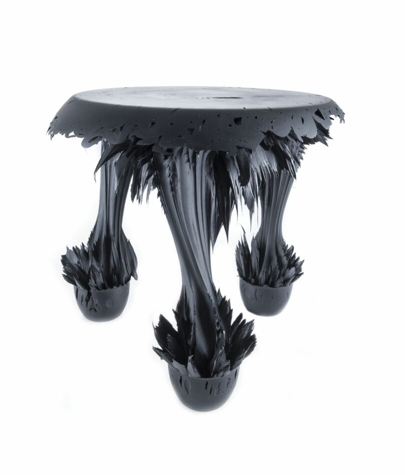 A sculptural table with a round top and three legs, appearing to be made from a dark, glossy material that has a melting or stretching texture
