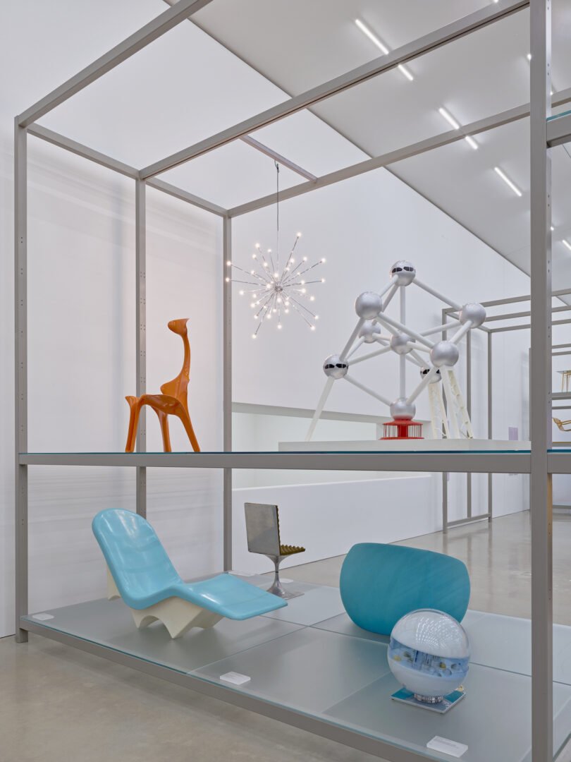 Display of various modern design objects set on metal shelving, including a blue chair, orange giraffe figure, small model of an atom-like structure, and a spherical lamp