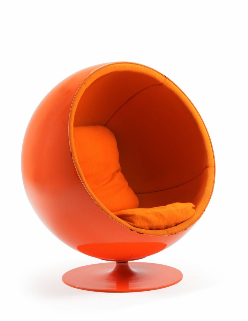 A modern, spherical orange chair with a cushioned interior and a swiveling base. The design resembles a partial globe with an open front