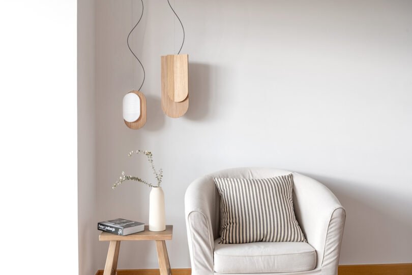 A minimalist living space with a white armchair, striped cushion, small wooden side table holding a vase with branches, and a book. Two wooden pendant lights hang on the white wall
