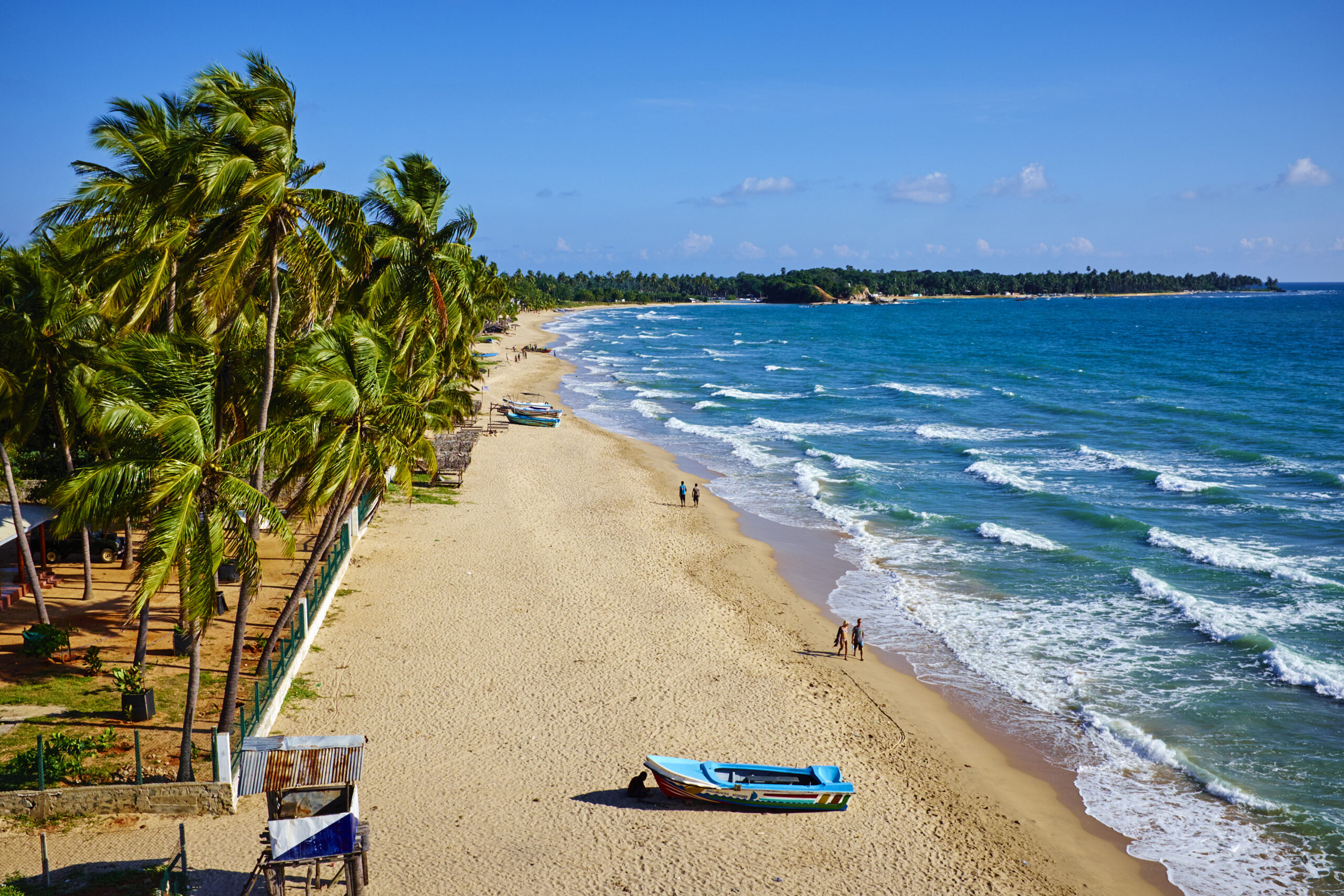 Uppuveli beach, Trincomalee, Sri Lanka A scenic aerial shot of a mostly empty beach with palm trees, golden sand and gentle turquoise waves, two couples walking by the water and a small boat on the sand in the foreground