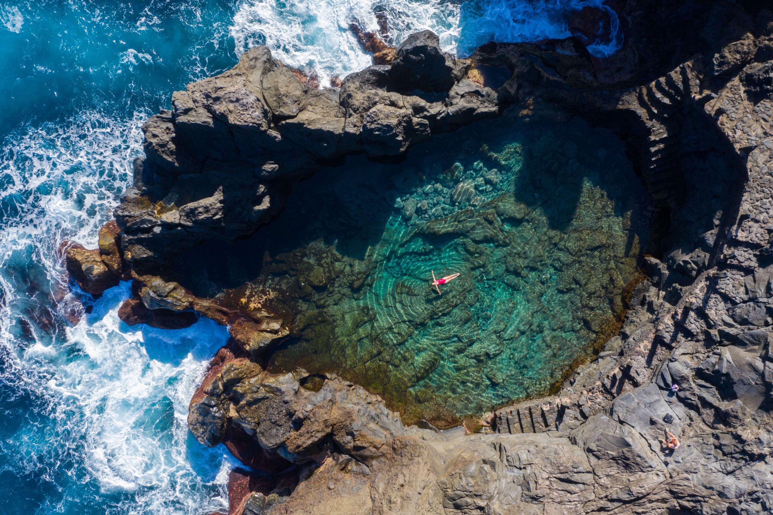 Tenerife A woman in a red bathing suit floats in a large rock pool next to the crashing surf