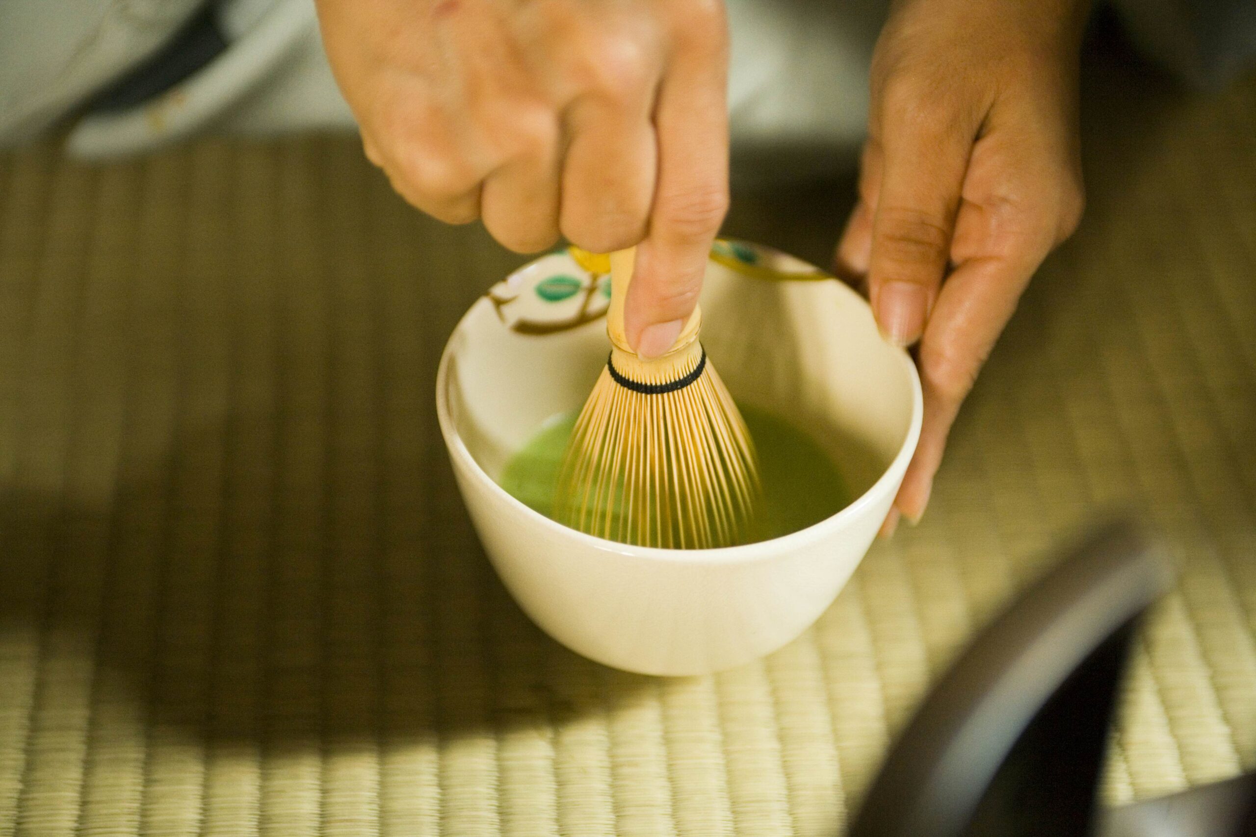 A hand with a whisk stirs matcha -- powdered green tea -- in a small white bowl on a woven mat